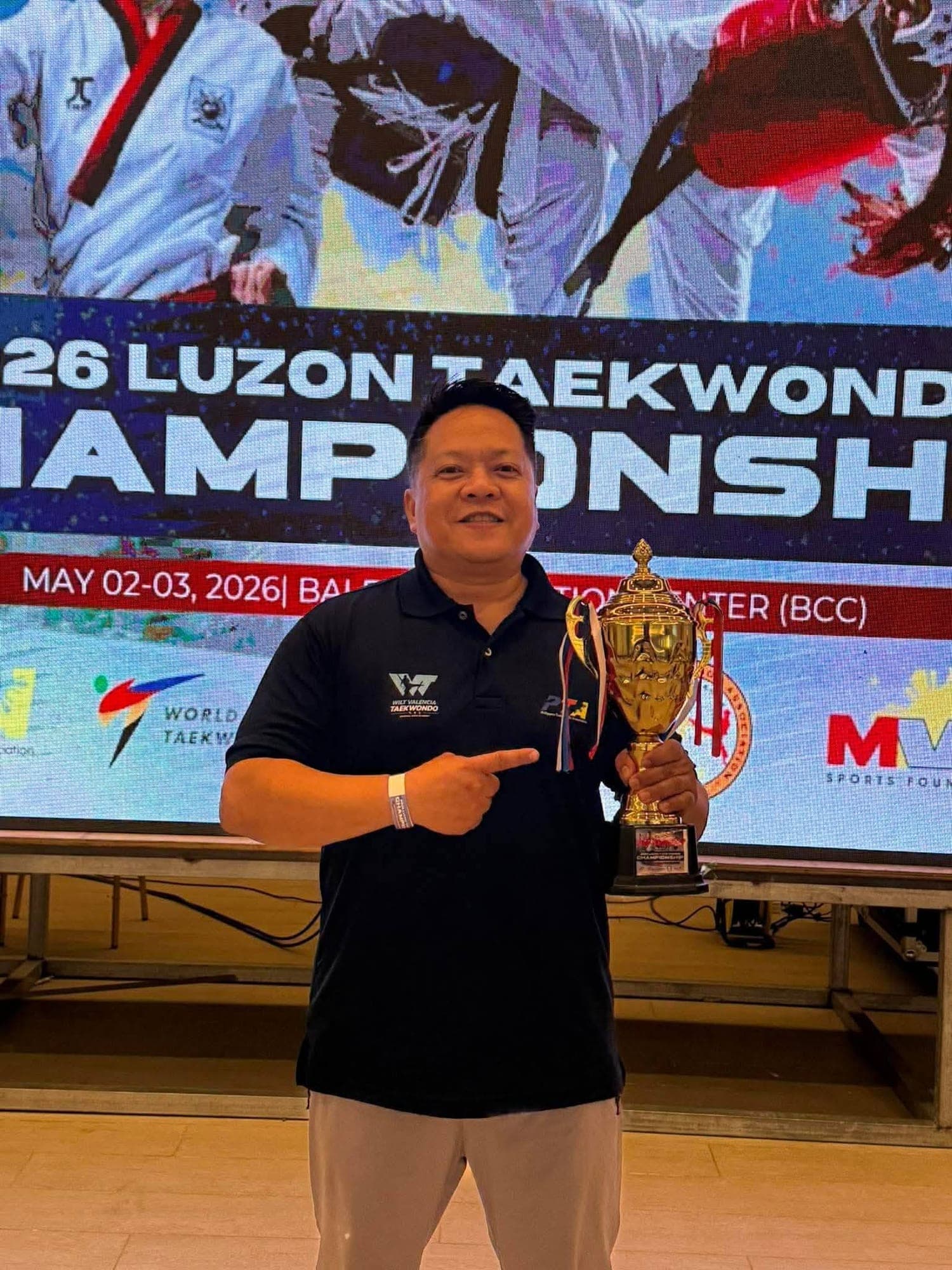Sabumnim Wilt Valencia holding a championship trophy in front of the 2026 Luzon Taekwondo Championships screen.
