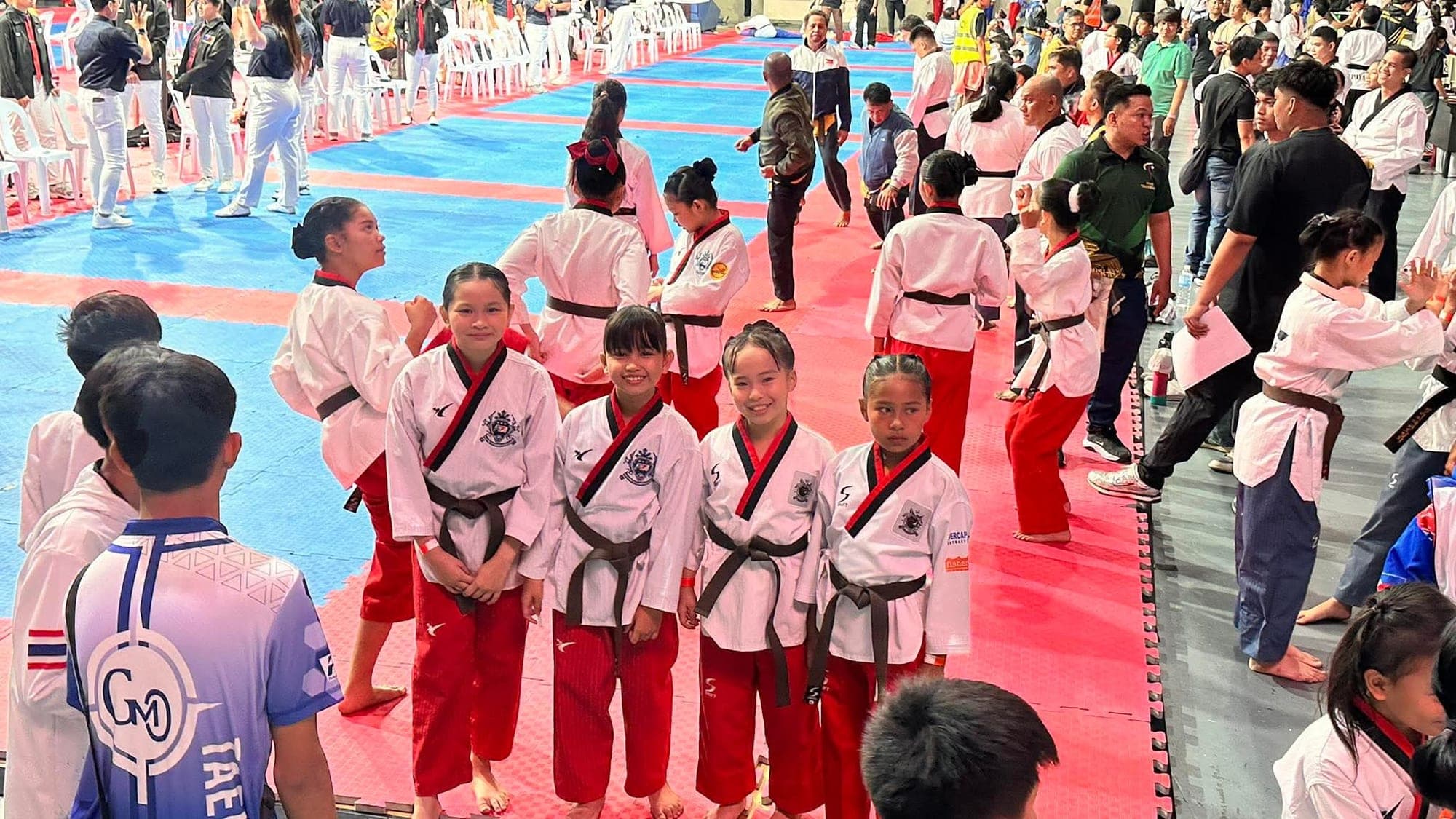 Four young WVT poomsae athletes in white and red uniforms with brown belts, smiling beside the competition mats.