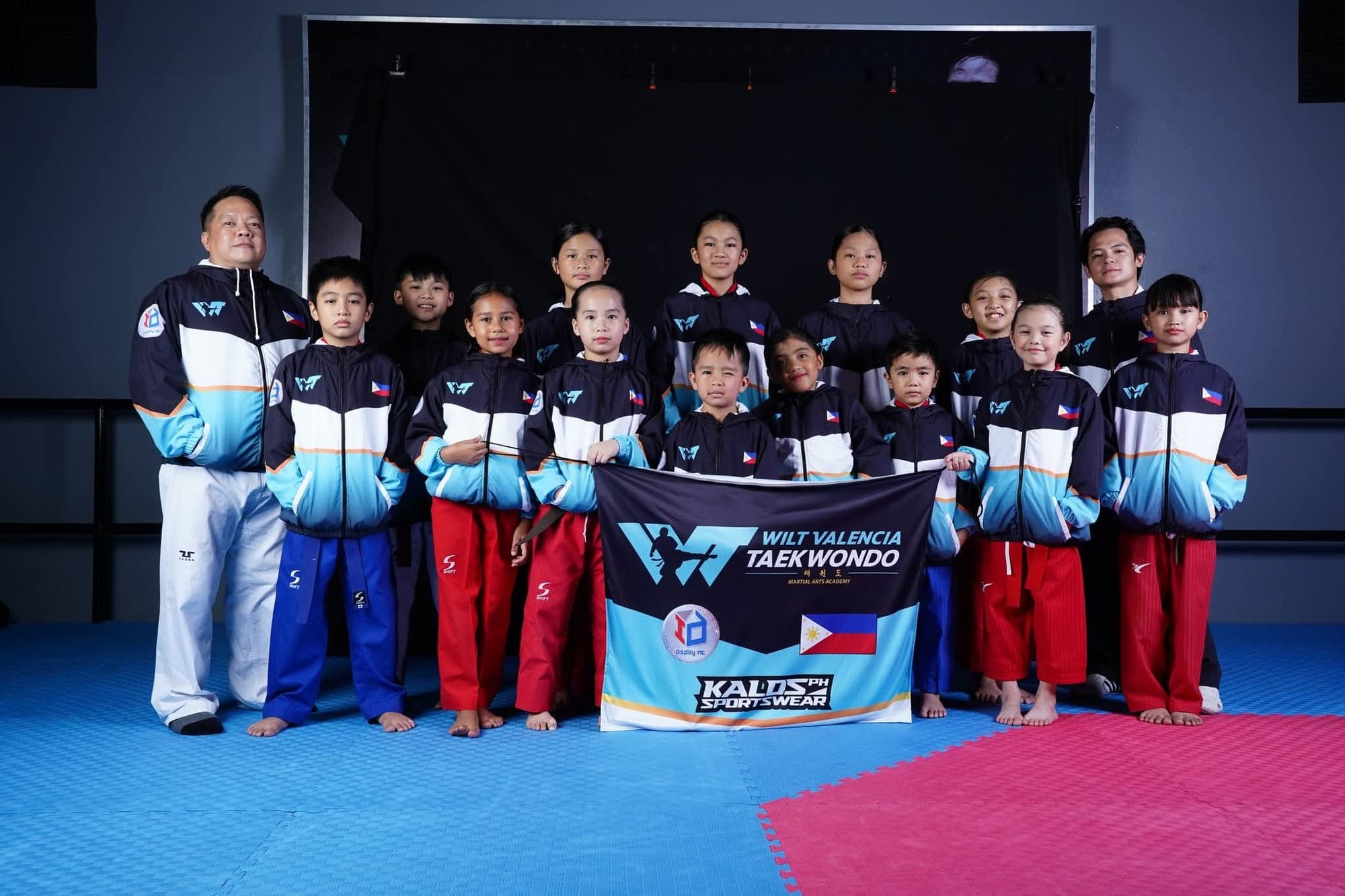 WVT athletes and coaches in matching team jackets standing on the mat behind the Wilt Valencia Taekwondo banner.